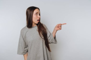 Uzun saçlı, gri, büyük tişörtlü genç bir kadın sürpriz yapıyor ve yan tarafı gösteriyor. Minimalist bir ortamda meşgul ve anlamlı görünüyor..