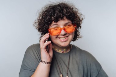 Kıvırcık saçlı neşeli bir insan canlı bir telefon görüşmesi yapar, şık turuncu güneş gözlüğü takar ve açık bir arkaplanda neşeli bir ifade sergiler..