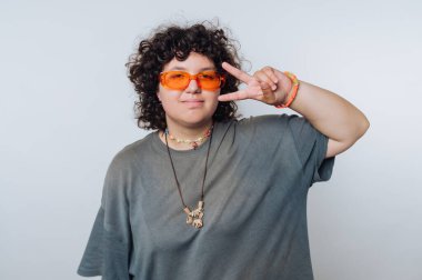 Kıvırcık saçlı genç bir yetişkin turuncu güneş gözlüğü ve rahat bir kıyafet giyiyor. Sade bir arka plana karşı durarak, özgüvenli ve kaygısız bir ruh sergileyerek bir barış işareti veriyorlar..