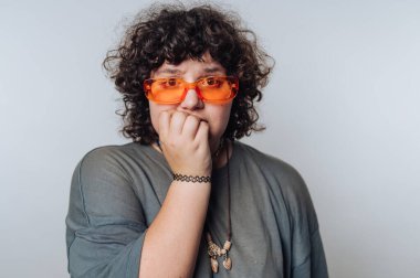 Genç bir yetişkin, kıvırcık saçlı ve kalın güneş gözlüklü gergin bir yüz ifadesini sergileyerek bir ışık arkaplanının önünde duruyor. Rahat kıyafet sıradan bir atmosferi işaret ediyor..