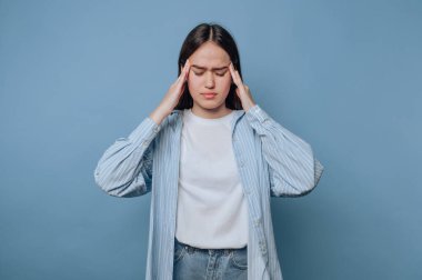 Genç bir kadın, elleri şakaklarının üzerinde duran, gözle görülür bir şekilde stresli, saf mavi bir zemine karşı duruyor. Günlük giysiler giyiyor ve rahatsızlık ifadesi sergiliyor..