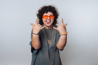 Kıvırcık saçlı bir genç, elleriyle neşeli bir jest yaparak coşku ve eğlenceyi ifade ediyor. Parlak güneş gözlüğü ve günlük giysiler takıyorlar. Kaygısız bir hava yayıyorlar..