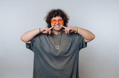 Büyük gri gömlekli ve parlak turuncu güneş gözlüklü bir kadın neşeli bir poz veriyor, parmaklarıyla barış işareti yapıyor ve neşeyle gülümsüyor..