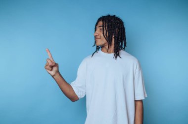 Saçları örgülü genç bir adam, yaratıcı ya da sıradan bir ortamda katıksız mavi bir arkaplana gülümserken şakacı bir şekilde parmağını gösterir. İfadesi coşku veriyor..
