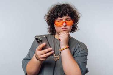Turuncu güneş gözlüğü takan kıvırcık saçlı genç, tarafsız bir arkaplanda derin derin düşünürken akıllı telefonlarına düşünceli bir şekilde bakar..