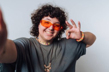 Kameraya neşeli bir ifadeyle poz veren, parlak bir stüdyo ortamında canlı turuncu güneş gözlüğü takan ve günlük kıyafet giyen, neşeli bir tavır sergileyen kişi..