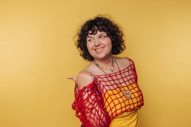 Kıvırcık saçlı genç bir kadın parlak bir kıyafet ve eşsiz kırmızı bir ağ bluz giyer. Parlak sarı bir arkaplan önünde neşeyle gülümser. Kendine güven ve kişilik yayar..