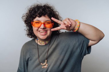 Kıvırcık saçlı neşeli bir genç, parlak turuncu gözlük, renkli bilezikler ve rahat bir tişört giyerek parmaklarıyla barış işareti yapıyor..