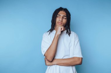 Genç bir birey, bir eli çenesinde, düşünceli bir ifade sergileyerek düşünceli bir şekilde ayakta duruyor. Arkaplan pürüzsüz bir mavi, sakin bir atmosfer yaratıyor..