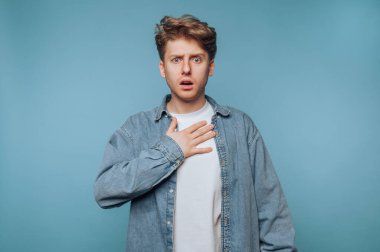 Beyaz bir tişörtün üstüne kot gömlek giymiş bir adam içeride duruyor. Şaşırmış görünüyor, elini göğsüne koyuyor. Arkaplan, onun şok ya da inançsızlık ifadesini artıran yatıştırıcı mavi bir renk..