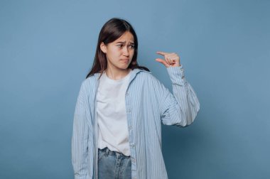 Açık renk çizgili gömlek ve kot pantolon giyen genç bir kadın mavi bir arka fonun önünde duruyor..