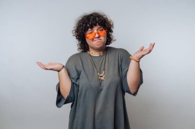 Genç bir birey omuz silkerek ayakta duruyor, şaşkın bir ifade sergiliyor. Rahat bir kıyafet ve parlak güneş gözlüğü takarak belirsizlik yaratıyorlar..