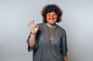 Kıvırcık saçlı neşeli bir birey turuncu güneş gözlüğü ve rahat bir kıyafet takar, el hareketleriyle poz verir ve kapalı bir ortamda gülümser. Parlak ve neşeli atmosfer havayı güçlendirir..