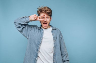 Genç bir adam mavi bir duvarın önünde duruyor, gülümsüyor ve elleriyle barış işareti yapıyor. Beyaz bir tişörtün üzerine kot ceket giyiyor, neşeli ve kaygısız bir hava yayıyor..