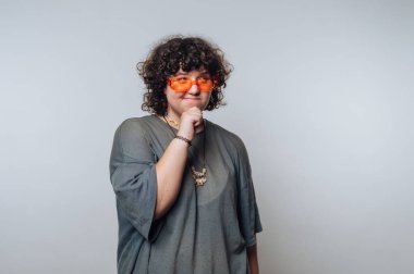 Genç bir yetişkin, kapalı alanda minimalist bir mekanda duruyor, turuncu güneş gözlüğü ve bol, rahat bir gömlek giyiyor. Çenelerine dokunurken neşeli bir ifadeyle düşünüyorlar..