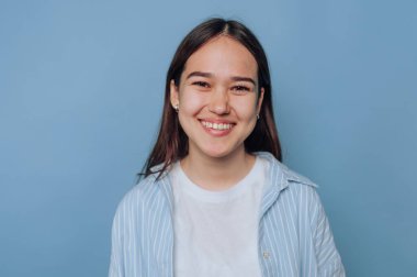 Genç bir kadın kendine güvenerek ayakta duruyor, açık mavi bir arka plana gülümsüyor. Neşeli yüz ifadesini tamamlayan, sıcaklığını ve yakınlığını arttıran sıradan bir kıyafet giyiyor..