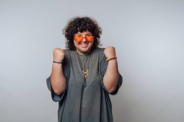 Kıvırcık saçlı genç bir kadın turuncu güneş gözlüğü takıyor ve kutlama için yumruklarını kaldırırken sevinçle gülümsüyor. Sıradan giyinmiş ve içeride harika vakit geçiriyor gibi görünüyor..