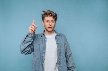 Genç bir adam bir parmağını açık mavi bir zemine kaldırmış, derin düşüncelere dalmış bir şekilde duruyor. O, sıradan bir kot ceket ve beyaz gömlek giyer, tefekkür eder..