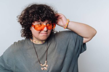 Kıvırcık saçlı biri, kendine has tarzını turuncu güneş gözlüğü ve renkli aksesuarlar takarak sergiliyor. Kapalı bir ortamda neşeli bir şekilde poz veriyorlar..
