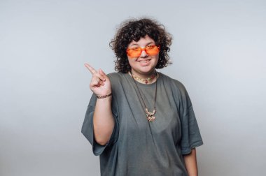 Kıvırcık saçlı ve parlak turuncu güneş gözlüklü genç bir birey büyük beden bir tişört giyerken şakacı bir şekilde işaret ediyor. Arka plan sade, neşeli ifadelerini ve tarzlarını vurguluyor..