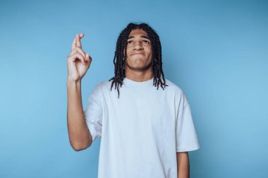 Genç bir birey umut dolu bir ifadeyle dimdik ayakta duruyor, parmaklarını çaprazlayarak, sağlam bir mavi arkaplanın önünde. Rahat duruş beklenti ya da iyi şanslar dilemeyi öneriyor..