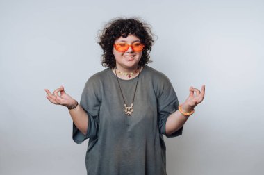 Genç bir kadın, kendine güvenen bir gülümsemeyle kıvırcık saçlarını ve turuncu güneş gözlüğünü gösteriyor. Minimum stüdyo arkaplanında elleri ile rahat bir jest yapıyor..