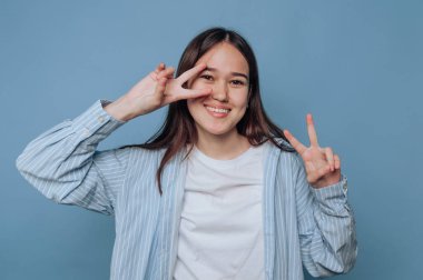 Genç bir kadın mavi bir arka plana karşı duruyor, gülümseyerek ve iki eliyle barış işareti yaparak, neşeli ve rahat bir tavır sergiliyor..