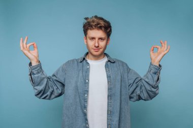 Genç bir adam farkındalık pratiği yapıyor, barış ve dengeyi el hareketleriyle gösteriyor. Sakin bir stüdyoda, dingin mavi bir arka planı var, sükuneti teşvik ediyor..