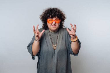 Kıvırcık saçlı biri canlı turuncu güneş gözlüğü takar ve kapalı mekanda anlamlı bir poz verir. Kıyafetleri modern moda estetiğini yansıtıyor, sanatsal bir hava yaratıyor..
