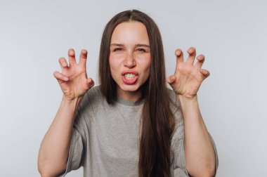 Uzun saçlı genç bir kadın elleriyle bir canavar jesti yapıyor. Gri bir gömlek giyiyor ve enerjik bir yüz ifadesi var, açık arkaplan üzerine kurulu..