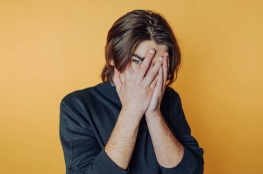 Genç bir adam parlak turuncu bir arkaplanın önünde duruyor, yüzünü ellerle örtüyor, canlı ve sanatsal bir ortamda utangaçlık ya da tefekkür duygularını yansıtıyor..