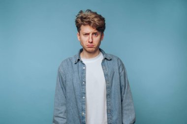 Genç bir adam beyaz bir tişörtün üzerine kot ceket giyerken ciddi bir yüz ifadesi sergiliyor. Yumuşak, mavi bir arkaplan karşısında durup duygusal bir anı yakalıyor..