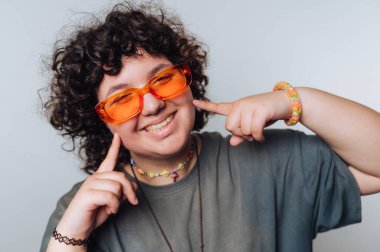 Kıvırcık saçlı biri parlak turuncu gözlük takarken parlak bir şekilde gülümser. Şakacı bir şekilde yanaklarını gösteriyorlar, sade bir arka plana karşı neşeli bir ifade sergiliyorlar..