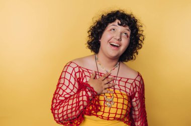Kıvırcık saçlı genç bir kadın parlak bir şekilde gülümsüyor, sarı bir elbisenin üzerine kırmızı ağlı bir bluz giyiyor. Mutlu ve meşgul görünüyor, neşeli bir ortamda pozitif enerji yayıyor..