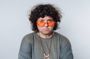 Kıvırcık saçlı, parlak turuncu güneş gözlüğü takan kişi rahat bir kıyafet giyerken kameraya dikkatle bakıyor. Renkli aksesuarlar görünüşlerine benzersiz bir dokunuş ekler..