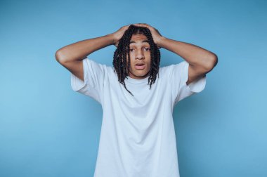 Genç bir adam sıradan beyaz bir tişört giyer, şaşırma ya da güvensizlik gösterir. Uzun örülmüş saçları ve çarpıcı bir kontrastlık yaratan mavi bir arka planı var..