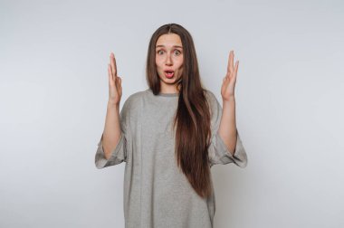 Uzun saçlı, sıradan gri gömlekli bir kadın içeride duruyor. Şaşırmış bir ifadesi var ve inanmamak için ellerini kaldırıyor. Arkaplan sade ve hafif.