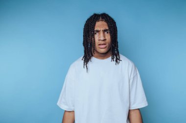 Örgülü saçlı bir genç, mavi bir arka plana karşı duruyor ve kafası karışık bir ifade sergiliyor. Ayarlar, canlı renklere sahip sıradan bir iç mekan fotoşopu olduğunu gösteriyor..