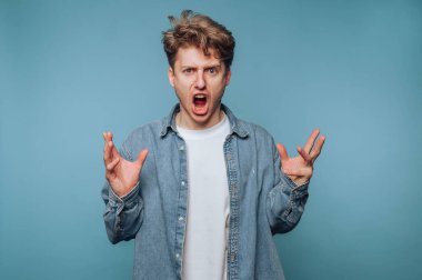 Genç bir adam mavi bir arka plana karşı duruyor, ellerini kaldırmış tutkuyla bağırıyor dramatik bir jestle. Günlük kıyafetleri ve yoğun ifadesi güçlü duygular taşıyor..