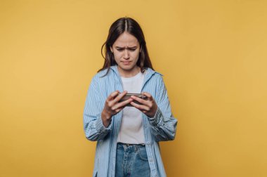 Parlak sarı bir duvarın önünde duran genç bir kadın, akıllı telefonuna odaklanırken hayal kırıklığına uğramış bir ifade sergiliyor, görünüşe göre bir meydan okumayla uğraşıyor..