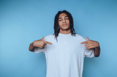 Saçı örgülü genç bir adam, sağlam bir mavi arkaplan önünde dikilirken kendinden emin bir şekilde kendini gösterir. Günlük beyaz tişörtü rahat ve havalı bir hava yaratıyor ve tarzını vurguluyor..