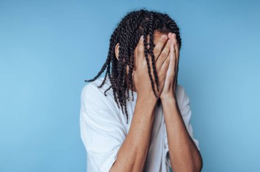 Uzun kıvırcık saçlı genç bir adam iki eliyle yüzünü kapatıyor. Kapalı bir ortamda, yumuşak mavi bir arka planda derin duygular aktarıyor..