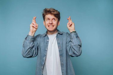 Kıvırcık saçlı ve kot ceketli genç bir adam açık mavi bir arka fonda duruyor, umut içinde gülümsüyor ve dua ediyor. İfadesi pozitiflik ve beklenti yayıyor..