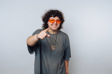 Kıvırcık saçlı ve turuncu güneş gözlüklü genç bir birey açık renkli bir stüdyoda duruyor, enerjik bir şekilde gülümsüyor ve işaret ediyor. Rahat kıyafetleri rahat bir hava veriyor..