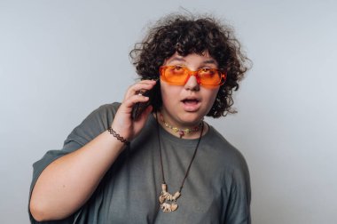 Kıvırcık saçlı biri telefonda konuşuyor, şaşırmış gibi bakıyor. Kapalı alandalar, turuncu güneş gözlüğü takıyorlar ve günlük kıyafetler giyiyorlar, eşsiz bir stili sergiliyorlar..