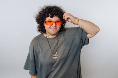 Kıvırcık saçlı ve turuncu güneş gözlüklü biri güvenli bir şekilde içeride duruyor, spor kıyafeti giyiyor. El yapımı aksesuarlar giyiyorlar ve bu parlak ortamda rahat bir hava katıyorlar..