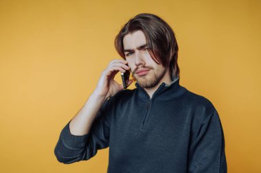 Uzun saçlı genç bir adam kulağına akıllı telefon dayıyor. Parlak sarı bir arkaplanda dururken yüzünde odaklanmış bir ifade var. Ciddi bir konuşma öneriyor..