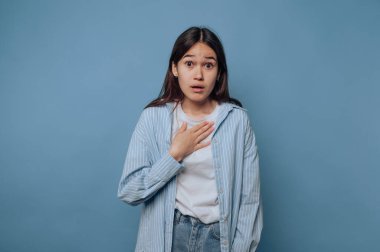 Genç kadın korkmuş görünüyor, elini göğsüne koyuyor. Açık mavi gömlek ve kottan oluşan günlük bir kıyafet giyer. Yumuşak mavi bir arka planda durur..