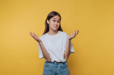 Şaşkın bir ifadeyle genç bir kadın omuz silkerek ellerini kaldırıyor. Sade bir beyaz gömlek ve kot şort giyiyor. Parlak sarı arka planına zıt düşüyor..