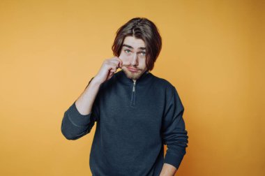 Koyu renk kazak giyen genç bir adam canlı turuncu bir duvarın önünde duruyor, saçlarıyla oynarken düşünceli bir ifadeyle doğrudan kameraya bakıyor..